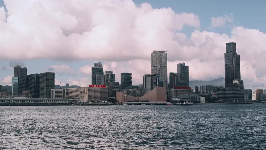 A wide static shot captures a dense cluster of tall skyscrapers and urban buildings along a waterfront viewed from a distance across a wide body of rippling water under a partially cloudy sky
