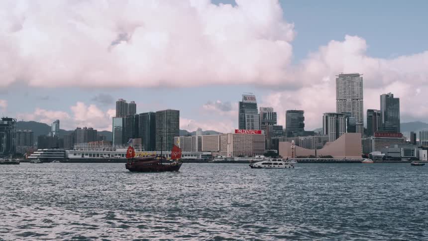 The video shows a wide shot of a bustling harbor with a modern city skyline in the background likely hong kongs victoria harboura traditional chinese junk boat with red sails is prominently featured sailing across the water in the for
