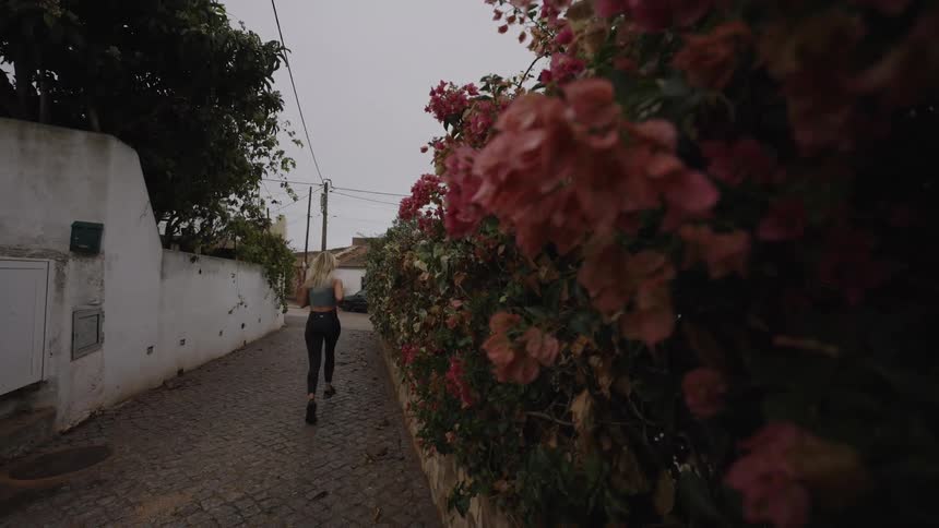 A side view shows a woman with blonde hair in a dark tank top and black leggings jogging away down a narrow cobblestone alley lined with a white wall and a dense hedge of pink bougainvillea flowers
