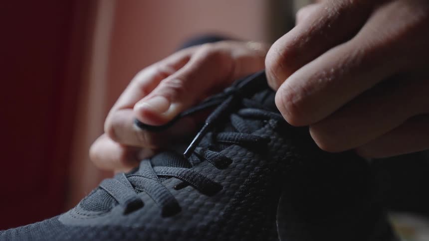 A pair of hands tightens the laces of a black textured athletic shoe in soft indoor light