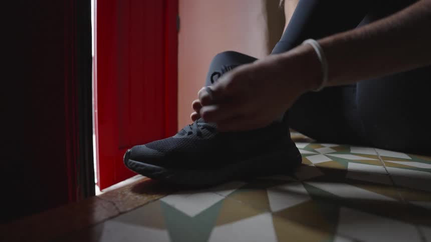 A persons hands tying the laces on a black athletic shoe while sitting on a patterned tile floor near a red door frame followed by the person standing up and walking out of the frame