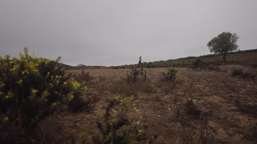 The video is an outdoor shot of a woman running along a desolate dusty hillside under an overcast sky the landscape is sparse with small shrubs and a single tree visible on the right side of the frame