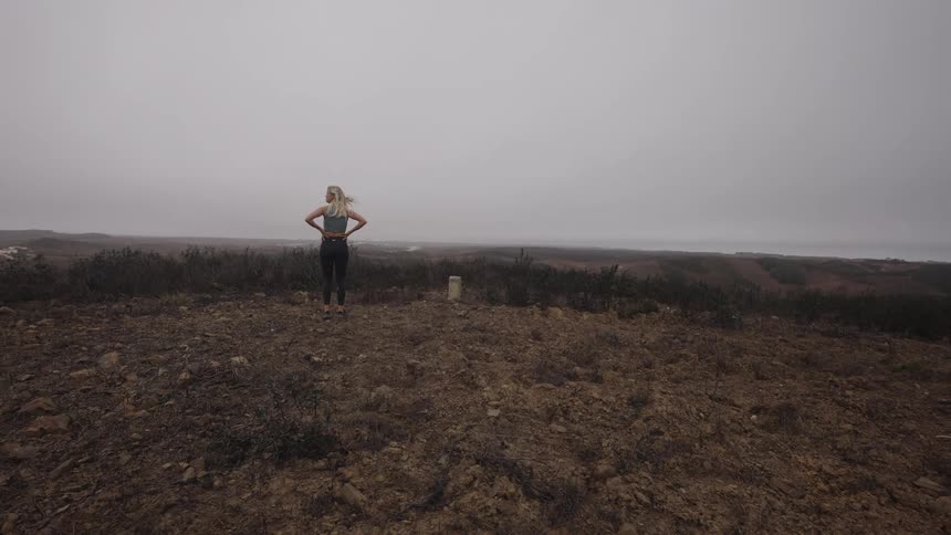 The video shows a blonde woman walking on a dry dirtcovered hill towards the ocean on a heavily overcast or foggy day she is wearing a teal or dark green top with black leggings and is viewed from behind providing a sense of scale against the