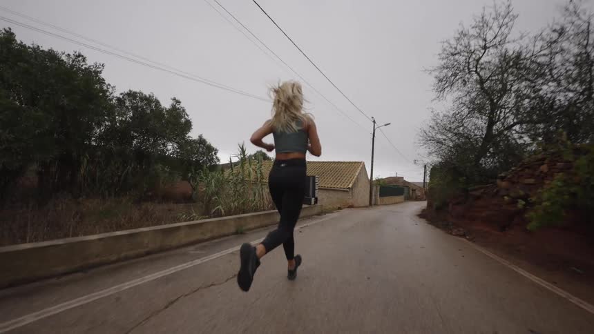 The video is a lowangle follow shot of a blonde woman running down a wet paved road she is wearing a grey sports top and black leggings running between rough stone walls and trees on an overcast day