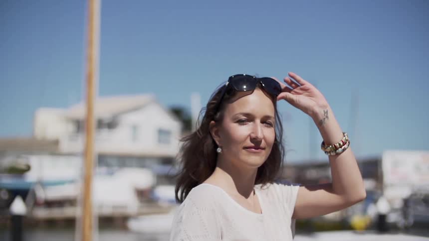 A young, brunette woman walking in front of docked boats while she turns, removes her sunglasses and smiles towards the camera