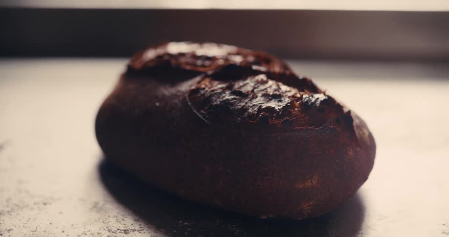 The video is a closeup shot of a single baked loaf of dark rustic bread resting on a metal tray the bread has a deep brown crackled crust and appears to be a sourdough or another artisan loaf with a soft focus on the background