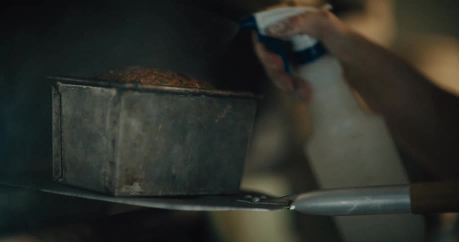 A square metal bread pan is pulled half way out of an oven, a baked loaf is visible with a brown crusty surface