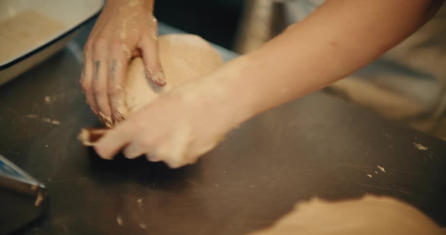 A close up shot of a baker's hands kneading and shaping dough on a stainless steel surface.