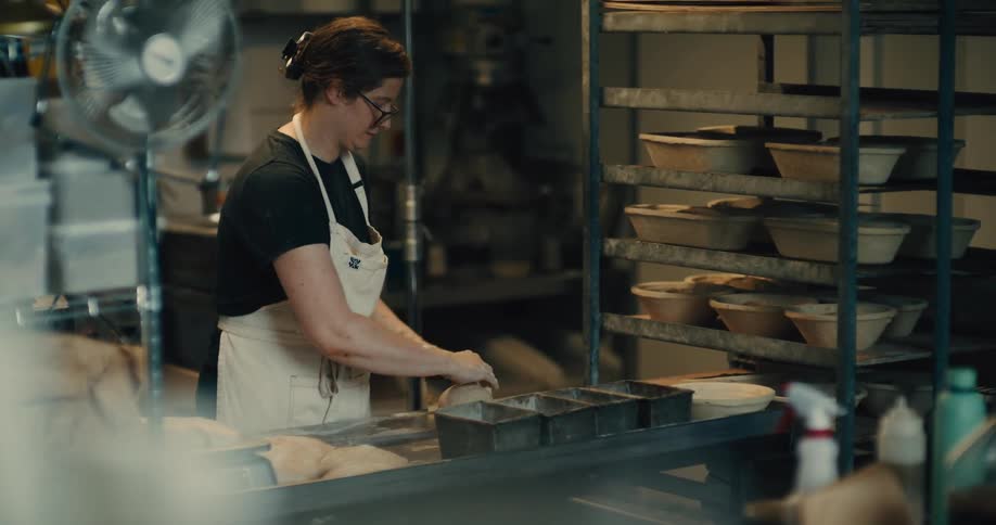 A baker wearing a white apron works with dough in an artisanal bakery, metal loaf tins are on the countertop