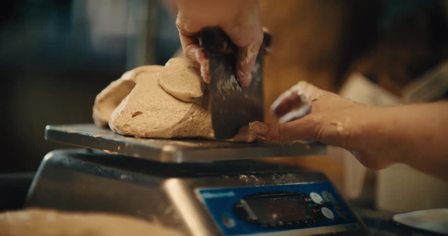 A baker weighs a piece of bread dough on a digital scale portioning it with a scraper