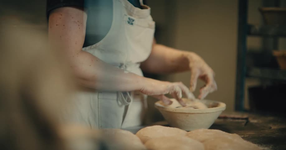 A medium close of a baker wearing a white apron placing a ball of dough into a baking dish, then sprinkling flower