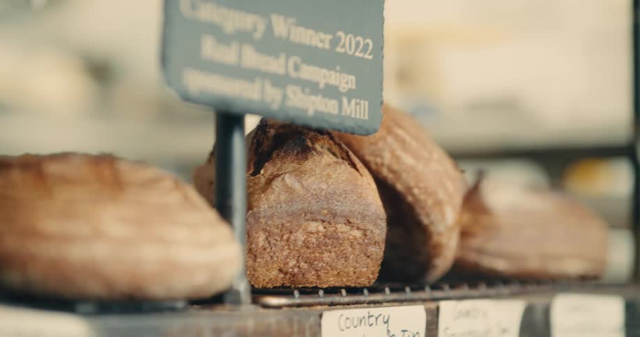 A close eye level view of several rustic sourdough loaves of bread sitting on a metal cooling rack on a bakery shelf next to a small blackboard sign in soft focus