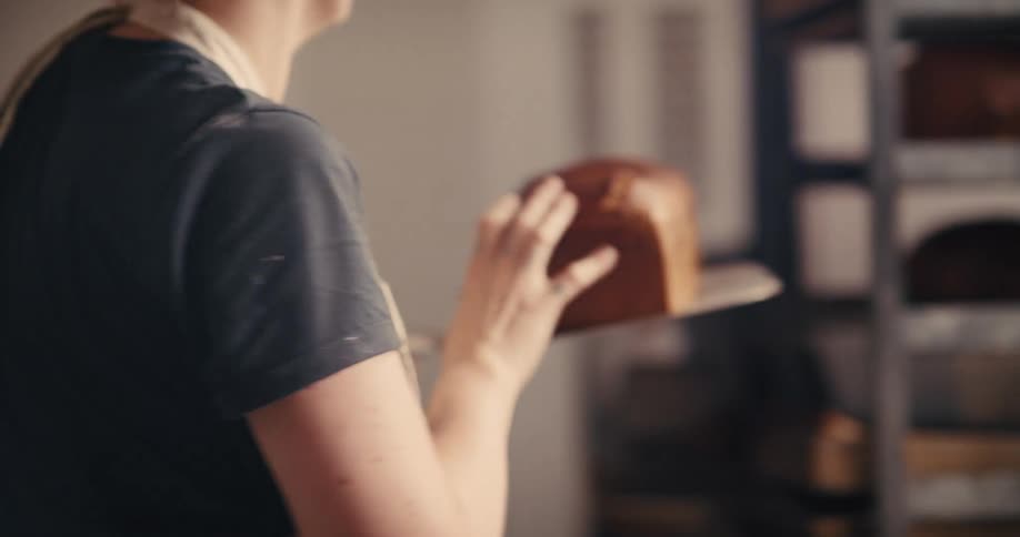 A female baker uses a paddle to slide loaves of bread out of a commercial oven and places them on a cooling rack