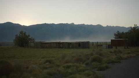 A man walks near a pen with some sheds on a ranch
