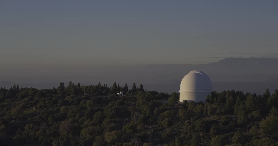 The video shows an establishing shot of an astronomical observatory situated on a forested mountain ridge a large white domed building characteristic of a telescope dome sits prominently on the hill surrounded by dark green trees against a pal