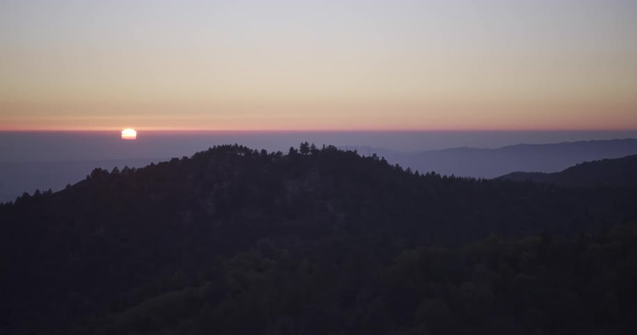 A distant wide view of a sunset shows a small orange sun dipping below a long dark forested ridge with hazy blue mountains layered in the background