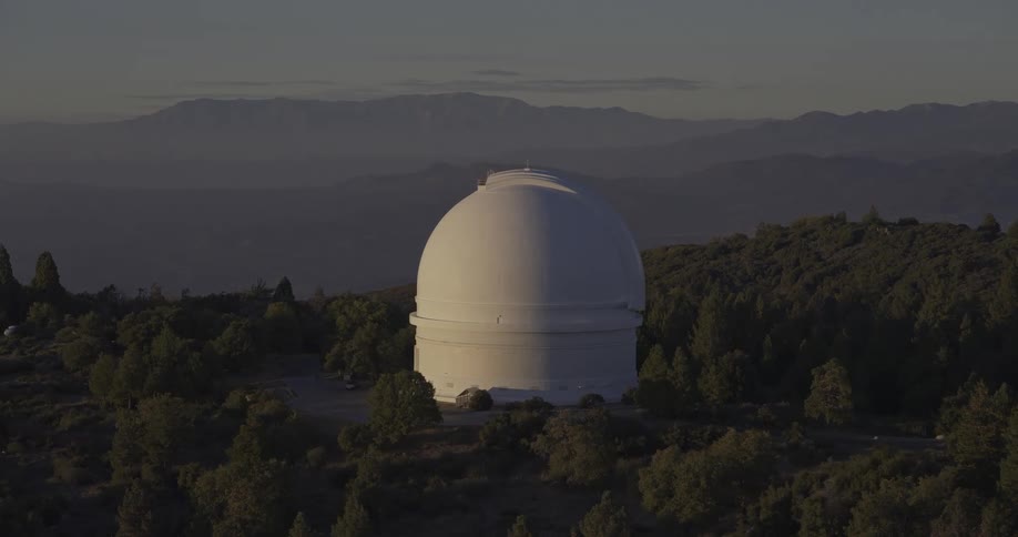 The video is an establishing shot of a large white astronomical observatory dome situated on a forested mountain ridge at dusk or dawn the observatory is surrounded by dark green trees with hazy layered mountains visible in the background agai