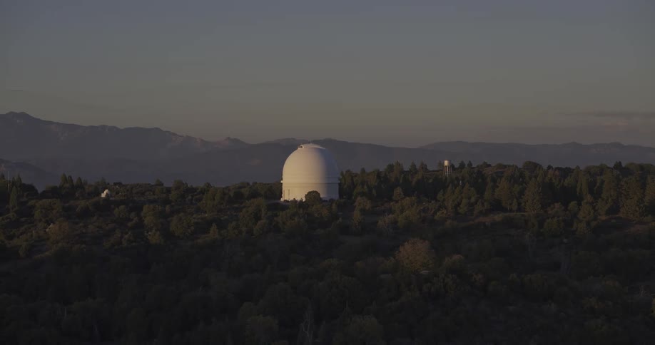 A white rounded observatory dome rising from a dense dark green forest on a hilltop with a layered mountain range visible in the deep background under a twilight sky
