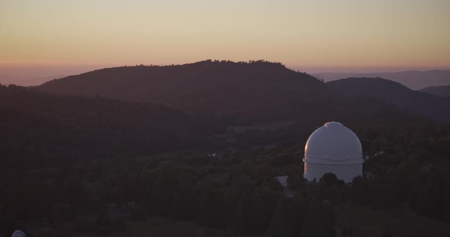 The video shows an aerial or wide shot of a white astronomical observatory dome on a densely forested mountain ridge the scene is at dusk or dawn with the sky fading from yelloworange to a deep blue above several layers of distant hazy mountai