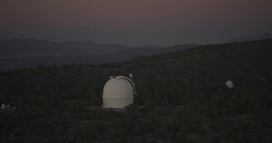 The video is an establishing shot of an astronomical observatory complex located on a dark forested hillside the large white telescope dome is prominent in the foreground with distant hazy mountains visible against the pale twilight sky