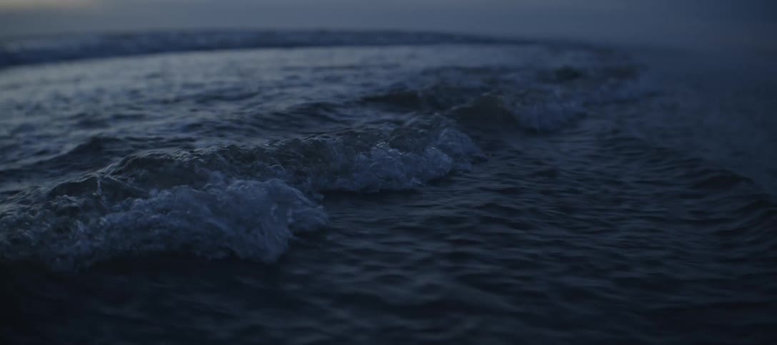 The video is a lowangle closeup shot of dark heavy ocean waves at dusk or nightthe waves roll and break towards the foreground creating white foam before receding back into the dark water