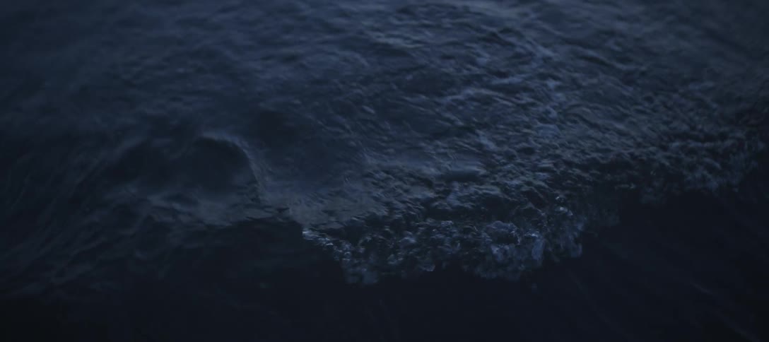 The video is a dark closeup shot of choppy deep blue or navy watera wave crests and breaks in the upper part of the frame then the water surface settles into continuous small ripples and movement