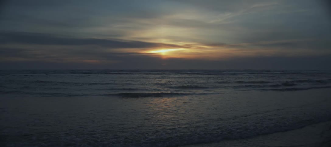 The video is an establishing shot of an ocean or sea under a cloudy sky at sunset or sunrise the sun is a bright yelloworange streak near the horizon shining through a dense layer of dark dramatic clouds