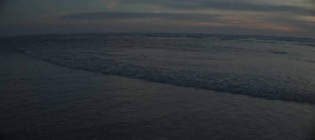 A high wide view of the dark ocean shows multiple lines of waves rolling onto the shore under a hazy dark sky with soft sunset colors on the horizon