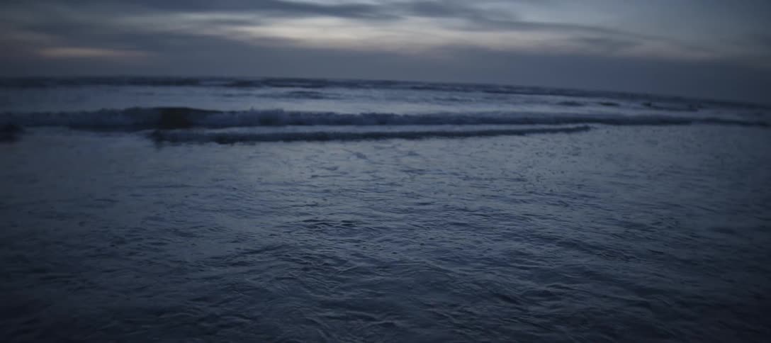A closeup of the dark churning water as a wave crests and breaks then rapidly tilts up and out to reveal the horizon where several lines of waves roll in beneath a cloudy muted sky