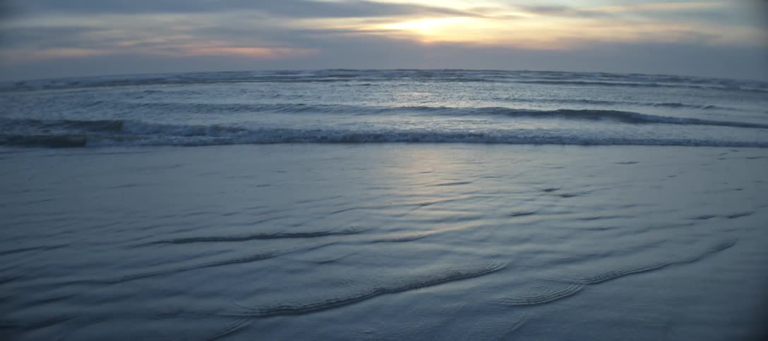 Water of the ocean rolling back and forth over the wet sand gradually tilting up to reveal a wider view of the rolling waves and the horizon with bright sunlight breaking through a band of dark clouds