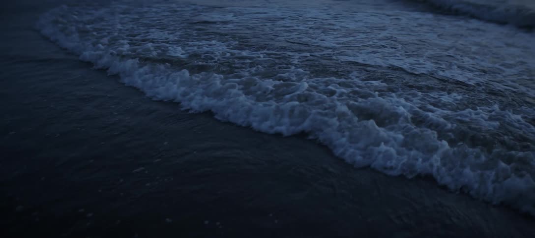 The dark blue surface of a body of water as a series of small waves crest and crash creating white foam that spreads and dissipates across the water and into the very dark foreground