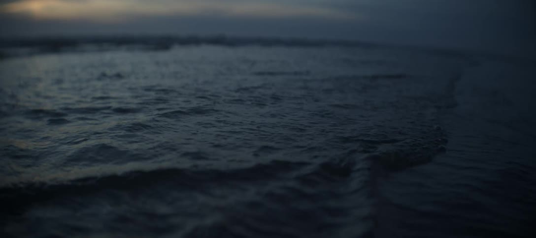 A very close low view of the dark deep blue ocean surface shows small waves continuously rolling toward the foreground with a soft sunset glow visible in the distant background sky