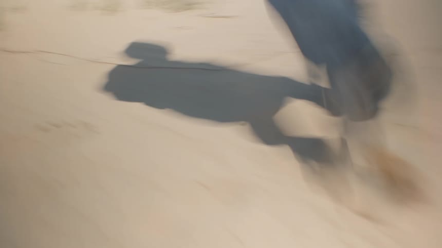 An abstract close lowangle view shows the blurred legs and shadow of a person walking across a light brown sandy surface with a few tufts of green grass