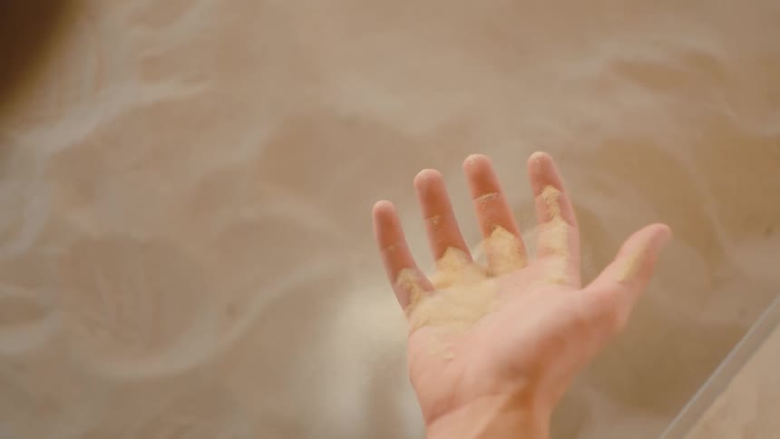 A close view of a persons hand sifting fine light beige sand that pours between the fingers in a flowing motion