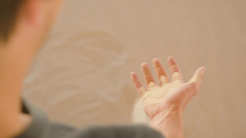 The video is a closeup shot of a persons hands as they sprinkle what appears to be dirt or seeds from one open palm onto the other the hands are held against a blurry warmtoned background possibly a wall or earth and the action suggests pla