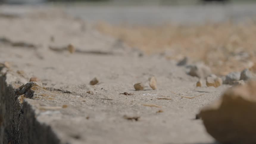 A flat gray surface with a sharp rough edge where small lightbrown irregular pebbles fall and bounce before settling on the dusty ground