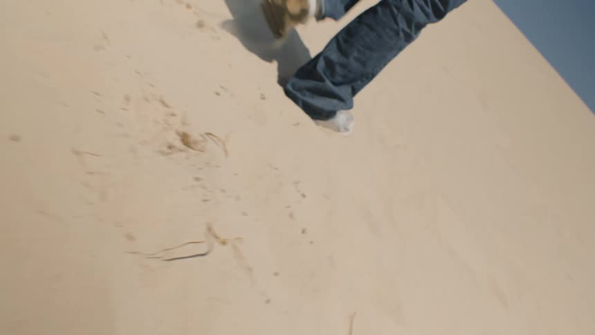 A lowangle view follows the legs of a person wearing blue jeans and white sneakers walking uphill on a steep dune of light tan sand under a clear blue sky
