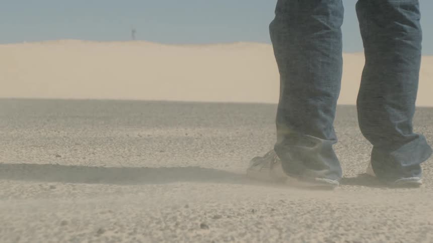 Lower legs and feet of a person standing still on a lightcolored granular ground as a low cloud of sand blows across the surface in a dry open outdoor environment with sand dunes in the background