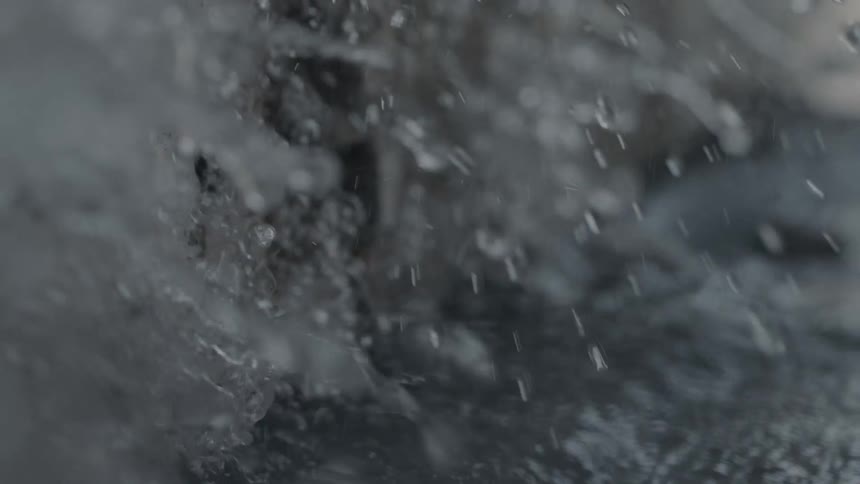 The video shows a closeup shot of a dark textured possibly cracked or decaying wall or stone surface and the ground next to itthen a sudden heavy stream or gush of water floods the frame splashing down onto the ground and the base of