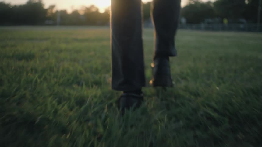 The video clip is a lowangle shot focused on a persons legs and feet walking across a bright grassy area at what appears to be sunset the individual is wearing dark pants and shoes and is walking from a fenced or gated area towards the camera