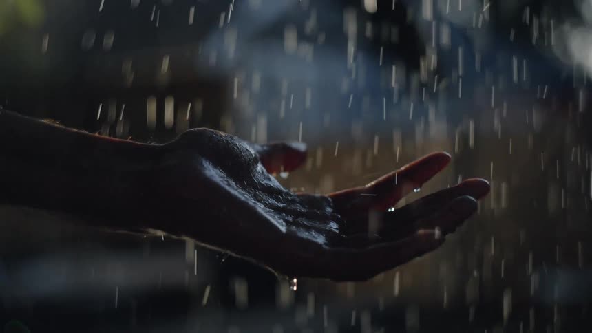 The video features a closeup dark silhouette of a persons outstretched hand catching falling waterheavy rain or droplets are clearly visible illuminated by a light source that highlights the water and the wet glossy hand