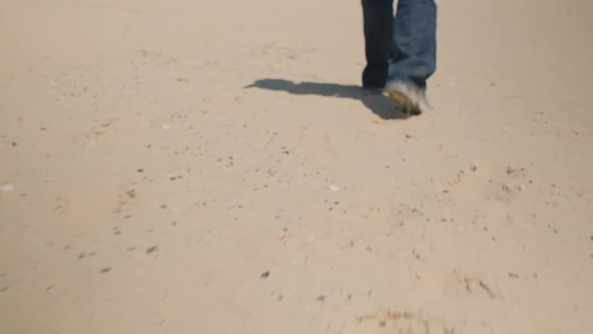 The lower half of a person wearing blue jeans and shoes as he walk away from the camera on a lighttan sandy surface while casting a long shadow behind him