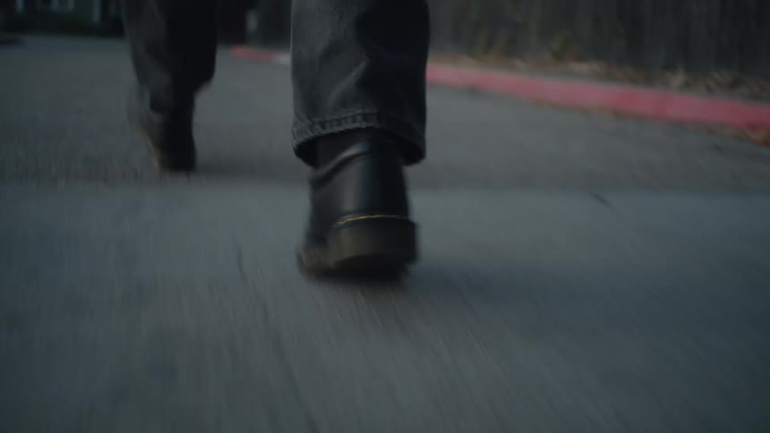 A close lowangle view follows the feet of a person wearing black boots and dark pants walking briskly down an asphalt residential street or sidewalk at dusk