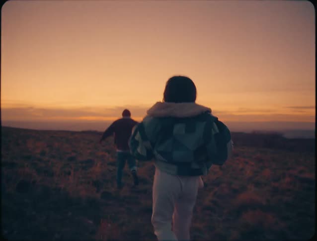 A couple runs playfully across a barren landscape at sunset with the young woman chasing the man in a lighthearted joyful way as golden light stretches across the open terrain