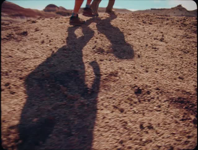 A closeup shot focuses on the lower legs and shadows of two people wearing boots as they move across a rough reddishbrown gravelcovered ground in a bright outdoor environment