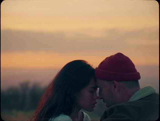 A romantic close view of a man wearing a red beanie and a woman with dark hair touching foreheads against a soft orange and gray sunset sky over a vast landscape