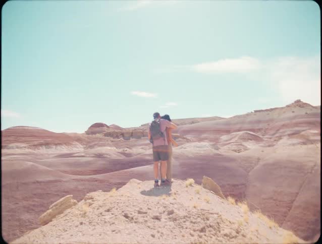 A person wearing a backpack standing on a rocky peak overlooking a vast multilayered arid landscape under a bright blue sky and then he hugs a person on the edge of a cliff