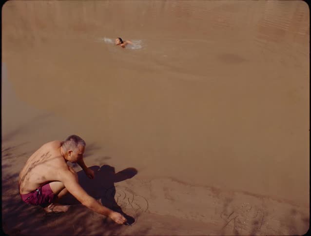 A barechested man in red shorts crouching at the edge of a large body of brown water and drawing with his finger in the wet sand while a woman swims past in the water behind him