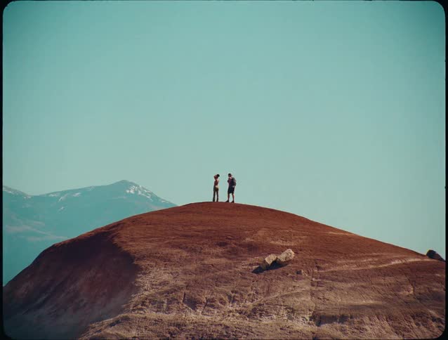 The video shows an expansive wide shot of a reddishbrown hill under a clear bright blue skytwo silhouetted figures stand small on the hills summit one raising their arms in triumph or joy with distant snowcapped mountains visible