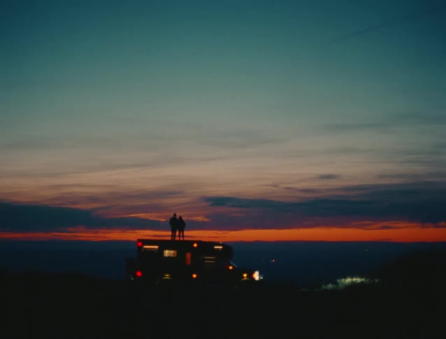 The video is a wide lowlight shot of two people standing as silhouettes on the roof of a van or motorhomethey are facing a dramatic sunset or sunrise which paints the vast sky in beautiful hues of orange red and deep blue